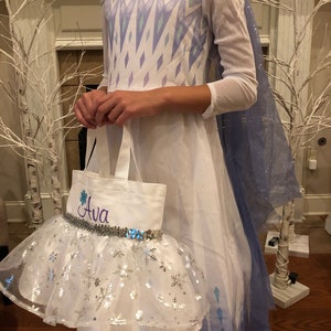 May include: A white dress with a blue and purple snowflake pattern, a blue cape, and a white tulle skirt with silver snowflakes. The dress has a white tote bag with the name "Ava" embroidered on it.