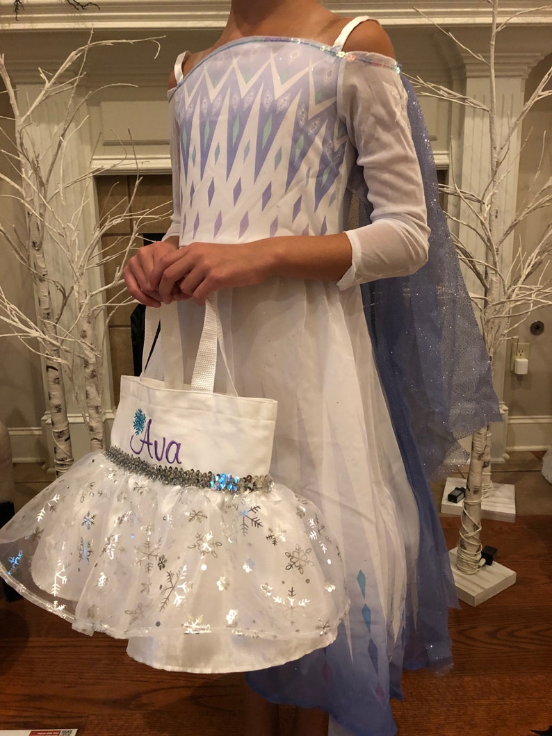 May include: A white dress with a blue and purple snowflake pattern, a blue cape, and a white tulle skirt with silver snowflakes. The dress has a white tote bag with the name "Ava" embroidered on it.