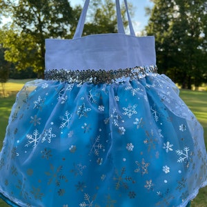 Puede incluir: Un bolso tote blanco con una parte inferior azul claro en forma de falda. La falda est&aacute; adornada con copos de nieve plateados y una banda de lentejuelas plateadas. El bolso tiene asas blancas y un ribete turquesa.