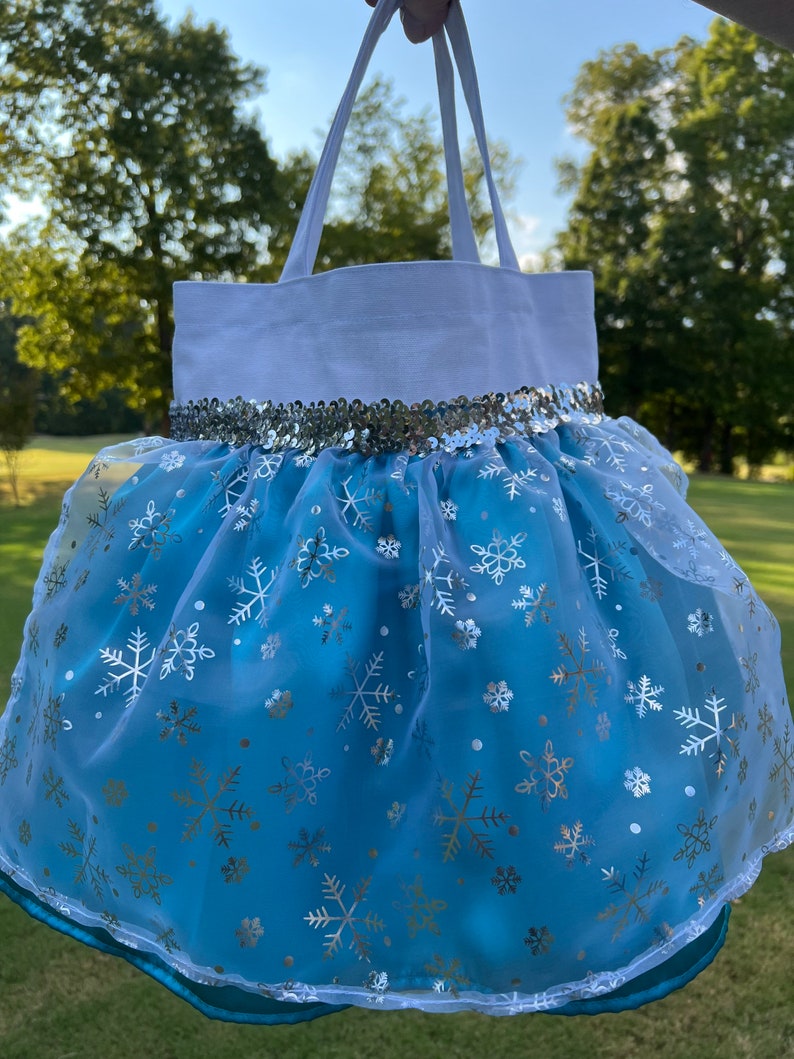 Puede incluir: Un bolso tote blanco con una parte inferior azul claro en forma de falda. La falda est&aacute; adornada con copos de nieve plateados y una banda de lentejuelas plateadas. El bolso tiene asas blancas y un ribete turquesa.