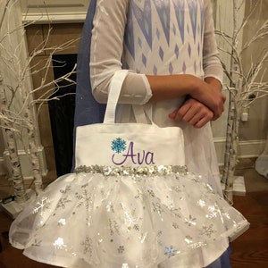 May include: White canvas tote bag with a purple embroidered snowflake and the name "Ava" in purple. The bag has a silver sequin trim and a white tulle skirt with silver snowflake accents.
