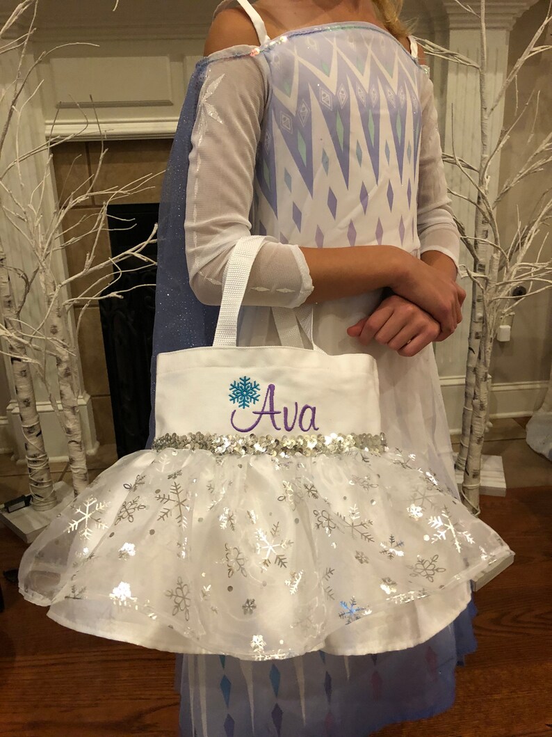 May include: White canvas tote bag with a purple embroidered snowflake and the name "Ava" in purple. The bag has a silver sequin trim and a white tulle skirt with silver snowflake accents.