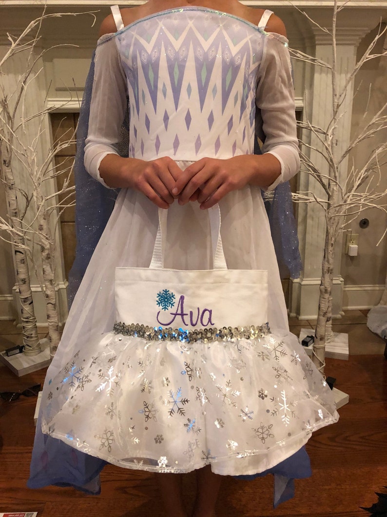 May include: White canvas tote bag with a silver sequin trim and a snowflake design. The bag is personalized with the name "Ava" in purple embroidery.