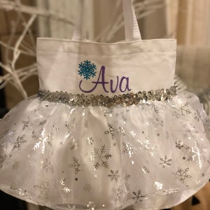 May include: White canvas trick-or-treat bag with a silver sequin trim and a tulle skirt with silver snowflake print. The bag is personalized with a blue snowflake and the name "Ava" in purple thread.