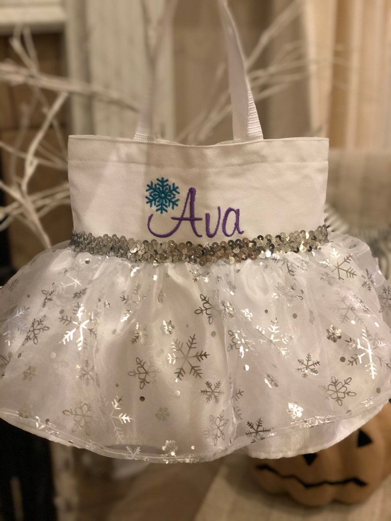May include: White canvas trick-or-treat bag with a silver sequin trim and a tulle skirt with silver snowflake print. The bag is personalized with a blue snowflake and the name "Ava" in purple thread.