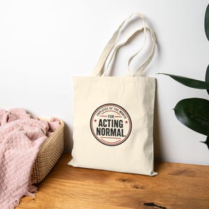 May include: A cream-colored canvas tote bag featuring a circular graphic with red and black text that reads "EMPLOYEE OF THE MONTH FOR ACTING NORMAL." The bag has two long shoulder straps and is made of a plain, light-colored fabric material.