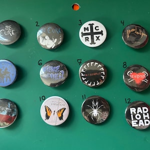 Può includere: Una collezione di dodici spille rotonde in metallo con vari disegni e nomi di band. Le spille sono argentate e nere, con testi come "The Cure", "My Chemical Romance" e "Radiohead". Sono disposte su una superficie verde.