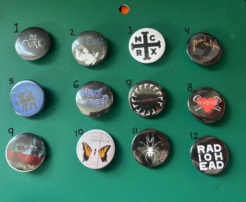 May include: A collection of twelve round metal pinback buttons with various designs and band names. The buttons are silver and black, with some featuring text like "The Cure," "My Chemical Romance," and "Radiohead." The buttons are displayed on a green surface.
