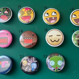 May include: A collection of twelve round pinback buttons with various cartoon and meme designs. The buttons feature colorful graphics, including faces with exaggerated expressions, a pixelated design, and a Nyan Cat image. The buttons are arranged on a green surface.