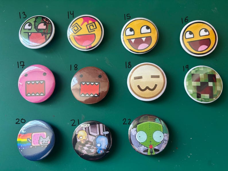 May include: A collection of twelve round pinback buttons with various cartoon and meme designs. The buttons feature colorful graphics, including faces with exaggerated expressions, a pixelated design, and a Nyan Cat image. The buttons are arranged on a green surface.