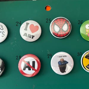 May include: A collection of eight round pinback buttons with various designs. The buttons feature text like "I <3 CATS", "I ART", and "I eat kids", along with images of Spider-Man, a frog, and a radiation symbol. The buttons are on a green surface.