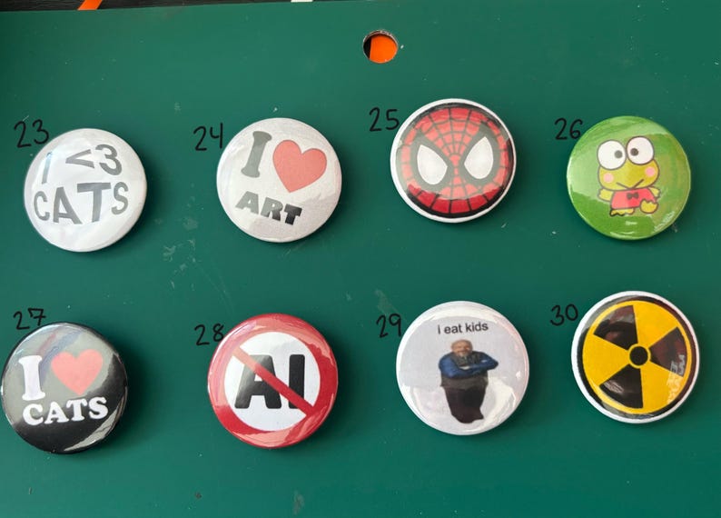 May include: A collection of eight round pinback buttons with various designs. The buttons feature text like "I <3 CATS", "I ART", and "I eat kids", along with images of Spider-Man, a frog, and a radiation symbol. The buttons are on a green surface.