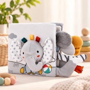 May include: A soft fabric baby book featuring a plush grey elephant with polka-dot ears on the cover. The book includes colourful ribbon tags, a printed beach ball graphic, and additional plush animal appendages extending from the side pages.