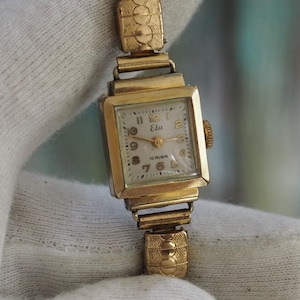 May include: A vintage gold-toned wristwatch with a rectangular face and a stretch band. The watch face has a light-colored background with black numerals and hands. The band and watch casing have a textured design.