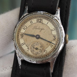 May include: Vintage wristwatch with a silver-toned case and a cream-colored face. The watch features black numerals and hands, with a smaller subdial. It is attached to a wide black leather strap.