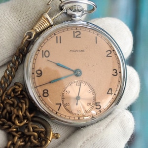 Könnte beinhalten: Eine Vintage-Taschenuhr mit silbernem Gehäuse, pfirsichfarbenem Zifferblatt und blauen Zeigern. Die Uhr hat ein kleines Sekunden-Zifferblatt und ist an einer goldfarbenen Kette befestigt. Die Uhr wird in einem weißen Handschuh gehalten.