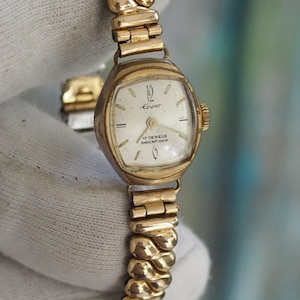 Könnte beinhalten: Eine Vintage-Armbanduhr in Goldoptik mit weißem Zifferblatt und goldenen Zeigern. Die Uhr hat ein rechteckiges Zifferblatt und ein flexibles, strukturiertes Armband. Das Zifferblatt zeigt den Markennamen "Lanco" und den Text "17 Jewels, Shockproof".
