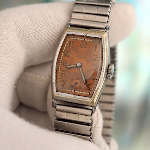 Könnte beinhalten: Vintage rechteckige Armbanduhr mit silberfarbenem Metallarmband und Gehäuse. Das Zifferblatt der Uhr ist in einem verblassten Orangeton gehalten und hat schwarze Ziffern und Zeiger. Die Uhr wird von einer behandschuhten Hand gehalten.