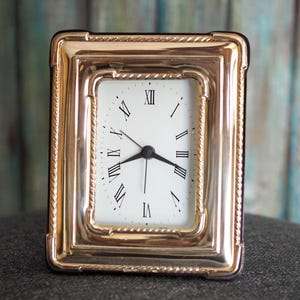 Può includere: Un orologio da scrivania rettangolare color oro con quadrante bianco e numeri romani neri. L'orologio ha lancette nere e una cornice color oro con un bordo a forma di corda. L'orologio è alto circa 10 cm.