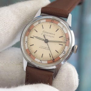 May include: Vintage Kampmann E. Heisingen wristwatch with a cream-colored face, featuring black numerals and hands, and a brown leather strap. The watch has a silver-toned case and a small crown. The face has a red and cream colored border.