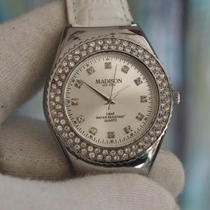 Könnte beinhalten: Eine silberfarbene Uhr mit weißem Lederarmband. Das Zifferblatt ist silberfarben mit rautenförmigen Stundenmarkierungen und einer Lünette, die mit kleinen, klaren Kristallen besetzt ist. Auf dem Zifferblatt steht "MADISON NEW YORK".