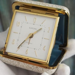Könnte beinhalten: Ein Vintage-Europa-Reisewecker. Die Uhr hat ein quadratisches weißes Zifferblatt mit goldenen Ziffern und Zeigern, eingebettet in einen goldenen Rahmen. Das Gehäuse ist cremefarben mit einem Blumenmuster und goldenen Zierleisten. Die Uhr ist geöffnet und zeigt ein blaues Innere.
