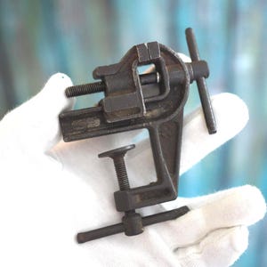 Könnte beinhalten: Ein kleiner, dunkelfarbener antiker Schraubstock mit Klemmfuß und Schraubmechanismus. Der Schraubstock hat einen Griff und eine Backe zum Halten von Gegenständen. Das Werkzeug ist aus Metall und scheint gut gebraucht zu sein.