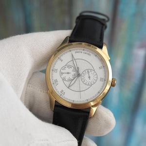 Könnte beinhalten: Eine goldfarbene Pierre Cardin Armbanduhr mit weißem Zifferblatt, schwarzem Lederarmband und römischen Ziffern. Die Uhr verfügt über Tages- und Datums-Hilfszifferblätter. Die Uhr wird von einer behandschuhten Hand gehalten.