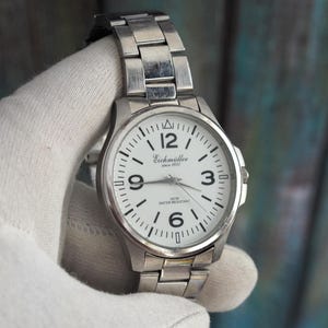 Könnte beinhalten: Eine silberfarbene Armbanduhr mit weißem Zifferblatt. Die Uhr hat schwarze Ziffern und Zeiger, mit dem Markennamen "Eichmüller" auf dem Zifferblatt. Das Armband besteht aus silberfarbenen Metallgliedern.