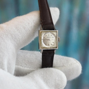 Könnte beinhalten: Eine Vintage Seiko Armbanduhr mit quadratischem silberfarbenem Gehäuse und hellem Zifferblatt. Die Uhr hat ein dunkelbraunes Lederarmband. Das Zifferblatt zeigt den Markennamen Seiko.