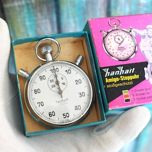 Könnte beinhalten: Eine Vintage Hanhart Amigo Stoppuhr in der Originalverpackung. Die Stoppuhr hat ein weißes Zifferblatt mit schwarzen Ziffern und eine Chromlünette. Die Box ist türkis und rosa mit den Worten "Amigo-Stoppuhr" und einem Läufer.