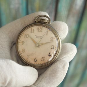 Könnte beinhalten: Vintage Westclox Pocket Ben Taschenuhr mit cremefarbenem Zifferblatt und schwarzen Ziffern. Die Uhr hat ein silberfarbenes Metallgehäuse und eine kleine Schlaufe oben. Die Zeiger sind goldfarben. Auf dem Zifferblatt steht "Westclox Pocket Ben".
