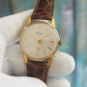 Könnte beinhalten: Eine goldfarbene Vintage-Raketa-Armbanduhr mit cremefarbenem Zifferblatt und goldenen Zeigern. Die Uhr hat ein braunes Lederarmband. Das Zifferblatt zeigt den Markennamen Raketa und den Text "16 Jewels".