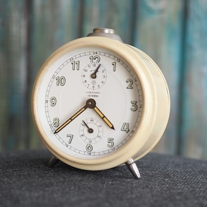 Könnte beinhalten: Cremefarbener Vintage-Wecker mit weißem Zifferblatt und schwarzen Ziffern. Die Uhr hat ein kleines Sekunden-Zifferblatt und die Marke "JUNGHANS -BIVOX-" ist sichtbar. Die Uhr hat ein Metallgehäuse und steht auf drei silbernen Beinen.