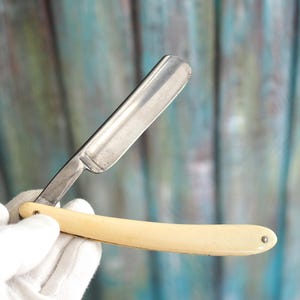 Può includere: Un rasoio diritto aperto con una lama argentata e un manico color crema. La lama è angolata e il manico è curvo. Il rasoio è tenuto contro uno sfondo sfocato.