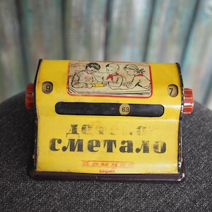 Könnte beinhalten: Vintage gelbes und braunes Metallspielzeug mit roten Knöpfen. Das Spielzeug hat ein Bild von Kindern und Text in einer Fremdsprache. Die Zahl 63 ist in einem kleinen Fenster sichtbar. Die Zahlen 9 und 7 befinden sich an den Seiten.