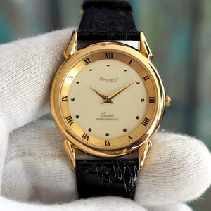 Könnte beinhalten: Eine goldfarbene Regent-Quarzuhr mit cremefarbenem Zifferblatt, schwarzen römischen Ziffern und einem schwarzen Lederarmband. Das Zifferblatt zeigt den Markennamen und die Worte "Quartz" und "water resistant".