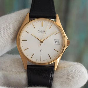 Op de afbeelding: Een goudkleurig Alpina sea-strong horloge met een witte wijzerplaat, gouden wijzers en een datumweergave op 3 uur. Het horloge heeft een zwarte leren band. De wijzerplaat heeft de tekst "ALPINA sea-strong".