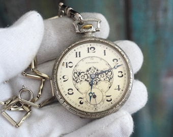 May include: Antique pocket watch with a cream-colored face, black numerals, and ornate floral detailing. The watch has a silver-tone case and a matching chain. The watch face reads "Mysteria".