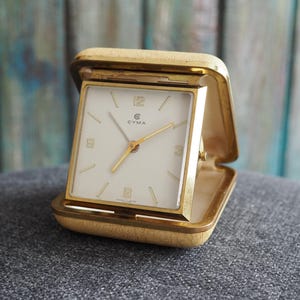 Könnte beinhalten: Ein goldfarbener Vintage-Reisewecker mit quadratischem, cremefarbenem Zifferblatt. Das Zifferblatt hat goldene Zeiger und Markierungen mit der Marke "CYMA". Die Uhr ist in einem scharnierten, strukturierten Goldgehäuse untergebracht. Maße: 7 cm x 7 cm.