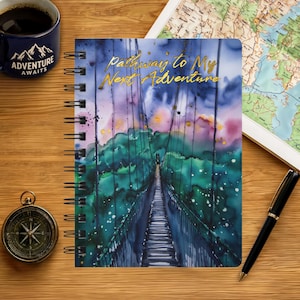 May include: A spiral-bound notebook featuring a watercolor illustration of a suspension bridge leading into a forest under a purple and blue sky. The gold text on the cover reads Pathway to My Next Adventure. The notebook is on a wooden surface.