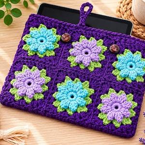 May include: A purple crocheted tablet sleeve featuring a floral granny square pattern with alternating light blue and lavender flowers on a green base. The sleeve includes a top loop for hanging and two dark brown circular buttons for closure.