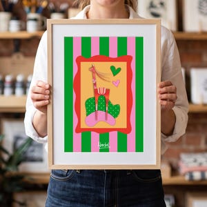 Puede incluir: Una lámina artística enmarcada con una ilustración estilizada de una jirafa sobre fondo amarillo, rodeada por un borde rojo festoneado. El fondo tiene rayas verticales verdes y rosas. Se ven un corazón verde y uno rosa. El texto Aberbel está impreso.