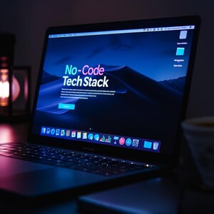 May include: A laptop displaying the words "No-Code Tech Stack" on the screen. The screen shows a blue and purple gradient background with a mountain range. The laptop is open on a dark surface.