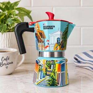 May include: A light blue stovetop espresso maker featuring a red lid and a black handle. The body is decorated with colorful illustrations of colonial architecture and palm trees, with the text "DESPIERTA BORICUA" printed near the top.