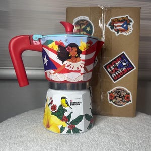 May include: A stovetop espresso maker featuring a red handle and lid, decorated with a Puerto Rican flag, a woman in a white dress, and yellow hibiscus flowers. The base displays a yellow bird, red berries, and the text Despierta Boricua.