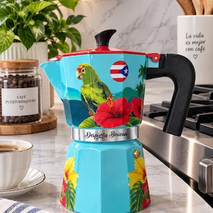 May include: A light blue stovetop espresso maker featuring a green parrot, red hibiscus flowers, a Puerto Rican flag emblem, and the text Despierta Boricua. The coffee pot has a red lid, a black handle, and yellow hibiscus floral accents.