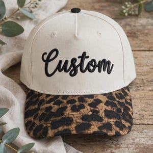 May include: A baseball cap with an off-white crown and a black button on top. The front features the word Custom embroidered in black script lettering. The brim displays a brown and black leopard print pattern with visible stitching lines.