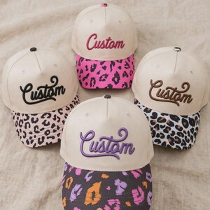 May include: Four baseball caps with cream-colored crowns and embroidered text reading Custom in various colors. Each cap features a unique animal print brim: pink and black, black and white, brown and black, and a multi-colored purple, orange, and black.