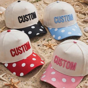 May include: Four baseball caps with off-white crowns and polka-dot brims. Each cap features the word CUSTOM in raised lettering. The brims and lettering are black, light blue, red, and pink, respectively, with matching top buttons.